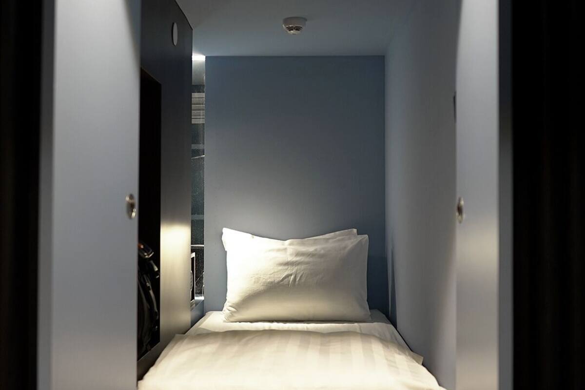 Ett kompakt hotellrum med en enkel säng, ett litet sängbord och en väggmonterad luftkonditioneringsanläggning.