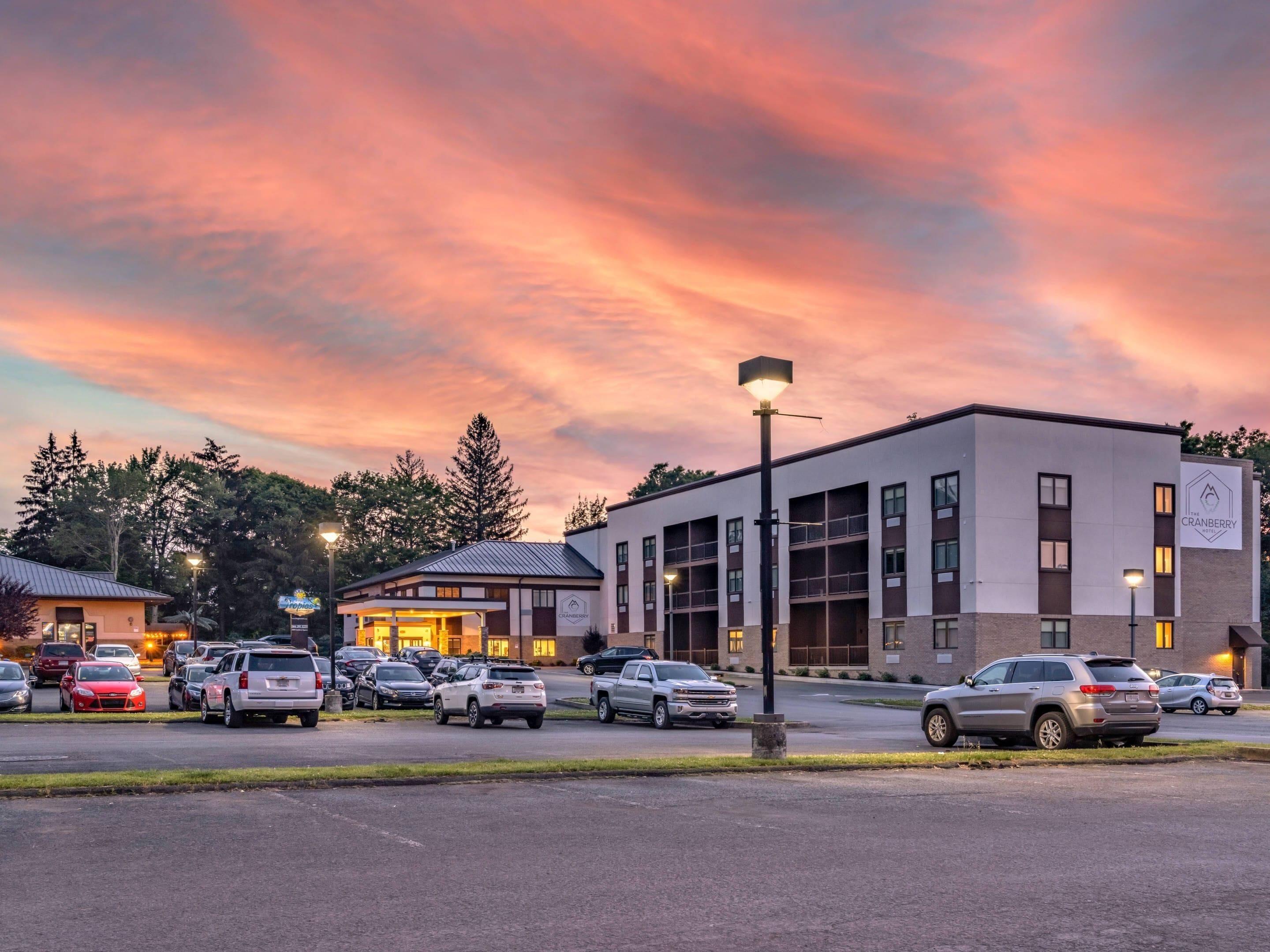Un hotel con diseño moderno, que cuenta con un estacionamiento con varios autos, un edificio de varios pisos y un cielo despejado con un atardecer.