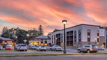 Un hotel con diseño moderno, que cuenta con un estacionamiento con varios autos, un edificio de varios pisos y un cielo despejado con un atardecer.