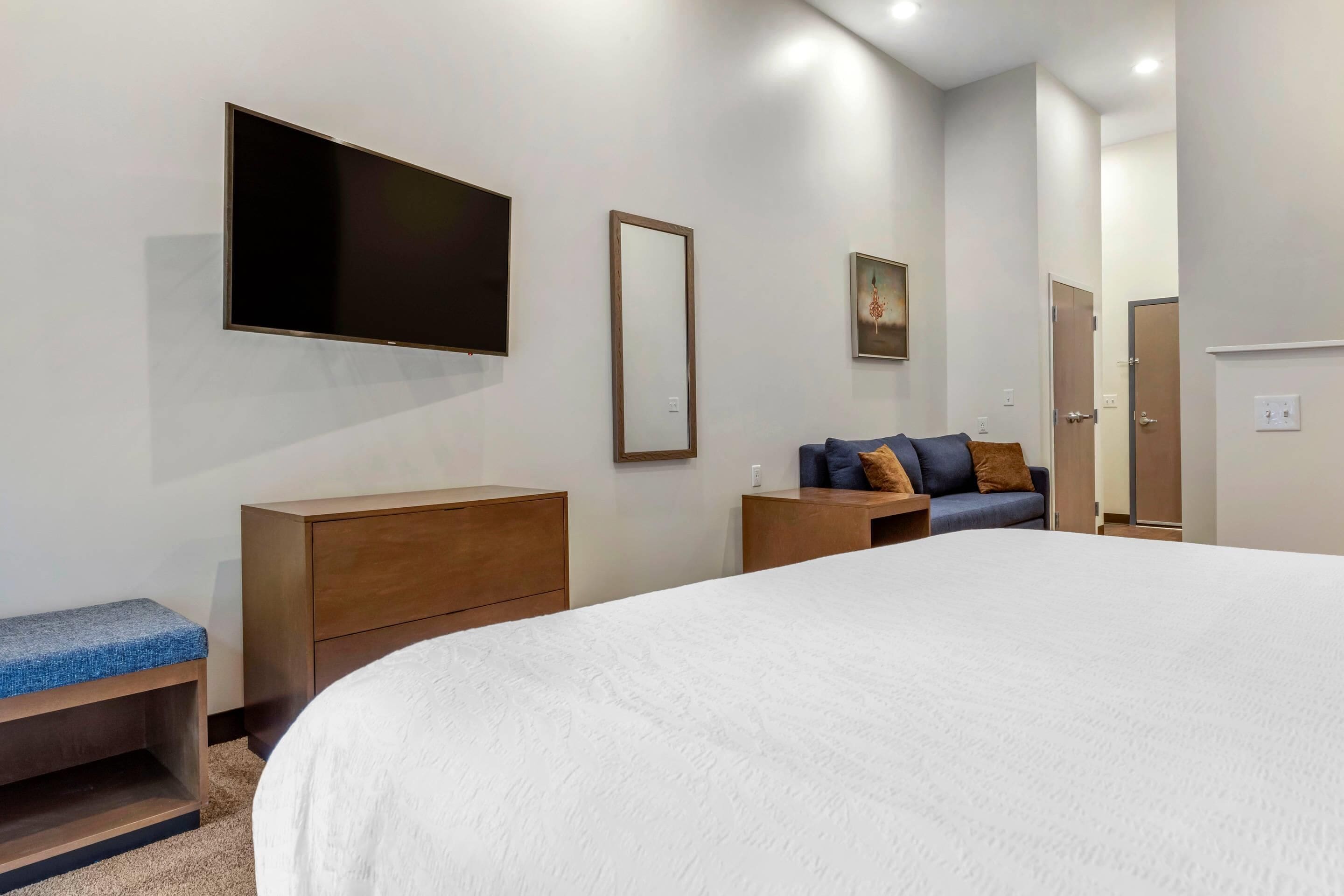 A modern hotel room with a large bed, a flat-screen TV mounted on the wall, a wooden nightstand, a bench, and a small sofa with cushions.