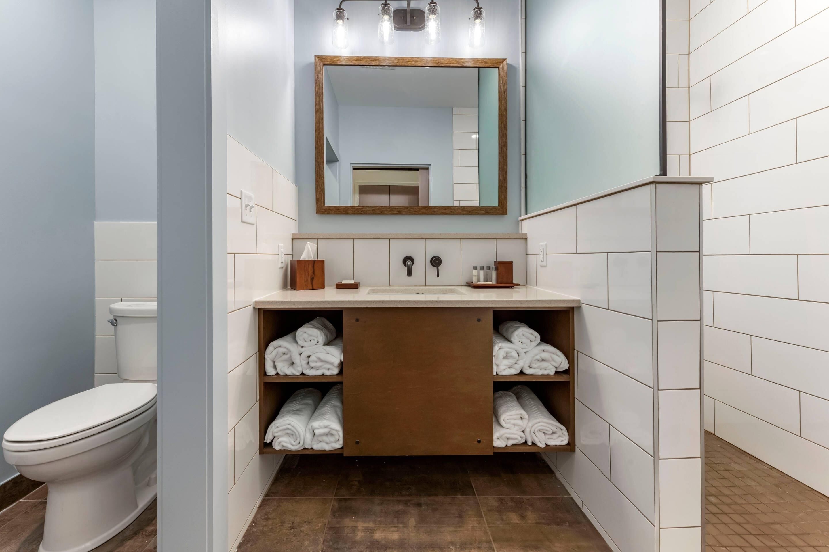 A modern bathroom with a white toilet, a wooden vanity with a mirror above it, and shelves with folded towels.