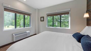 Un dormitorio con una cama grande, dos ventanas, un calentador montado en la pared y una mesita de noche con una lámpara.