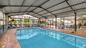 An indoor swimming pool with a covered roof, lounge chairs, and a patio area with tables and chairs.