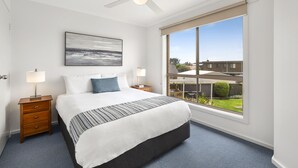 A bedroom with a large bed, a ceiling fan, a window with a view of the outside, and a nightstand with a lamp.