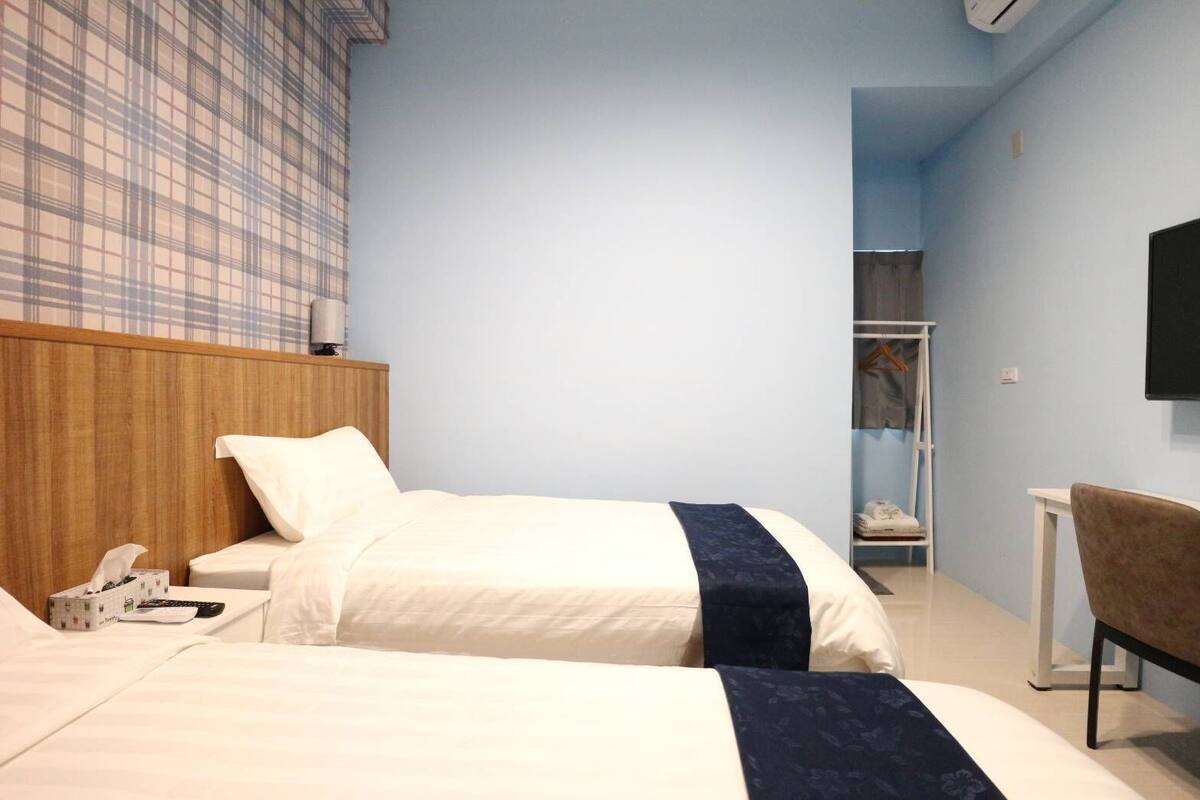 Habitación de hotel con dos camas, un cabecero de madera, una mesita de noche con lámpara, un armario y un televisor montado en la pared.