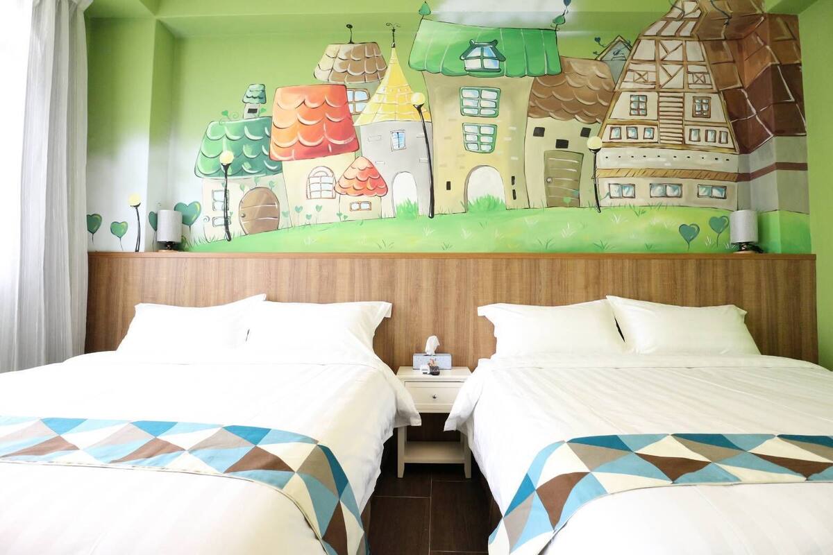 Habitación de hotel con dos camas, cabecera de madera, mesita de noche con lámpara y un mural de casas en la pared.