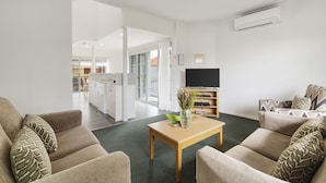 A modern living room with a sofa set, a coffee table with a vase of flowers, a flat-screen TV, and a kitchen in the background. - Whitecliffs Beach Resort (Rye)