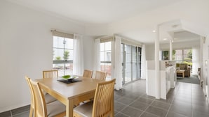 A spacious dining area with a wooden table and chairs, a kitchen in the background, and large windows with curtains. - Whitecliffs Beach Resort (Rye)