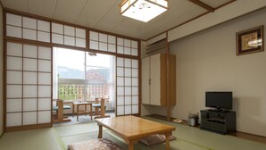 Perinteinen japanilainen huone, jossa on tatamimatto, matala puupöytä, televisiotaso ja liukuovet, jotka johtavat parvekkeelle, jolta avautuu näkymä vuorille.