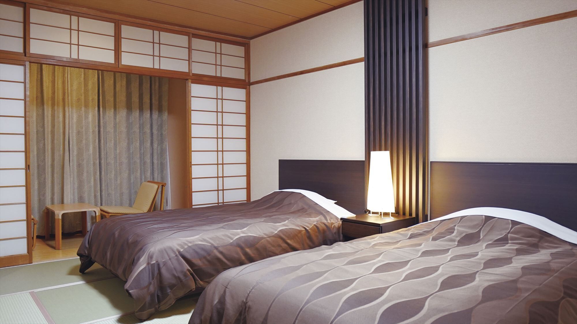 A traditional Japanese-style room with tatami flooring, two beds with patterned bedding, a wooden desk, and a sliding shoji screen.