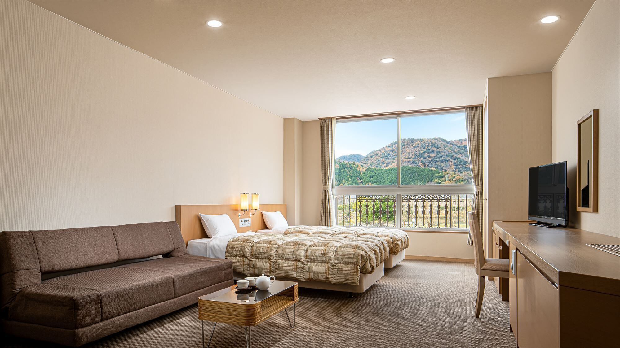 A hotel room with a bed, a sofa, a desk with a TV, and a balcony with a mountain view.