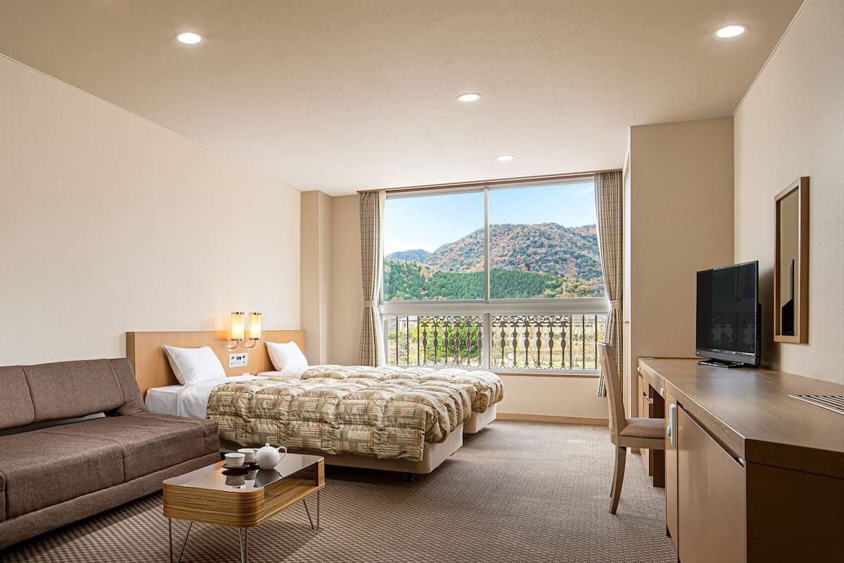 A hotel room with a bed, a sofa, a desk with a TV, and a balcony with a mountain view.