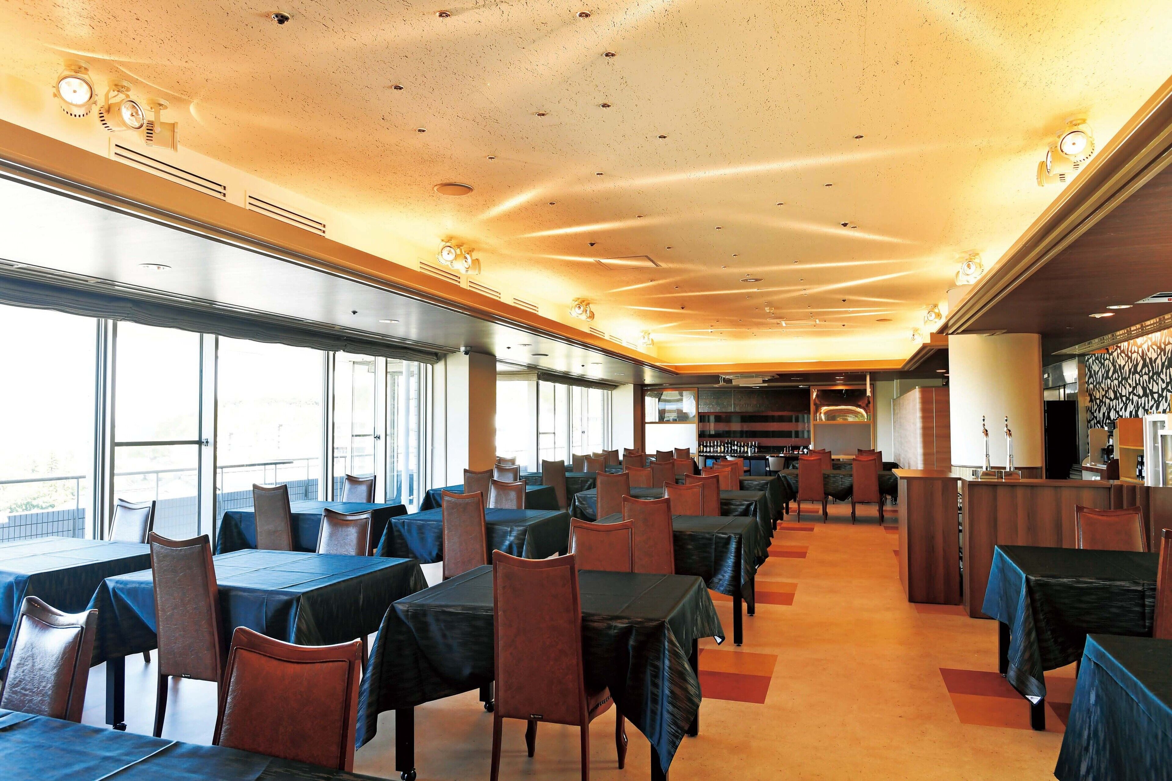 Un'ampia sala da pranzo con tavoli e sedie disposti in file, ampie finestre su un lato e un'area bar con bancone e sgabelli.