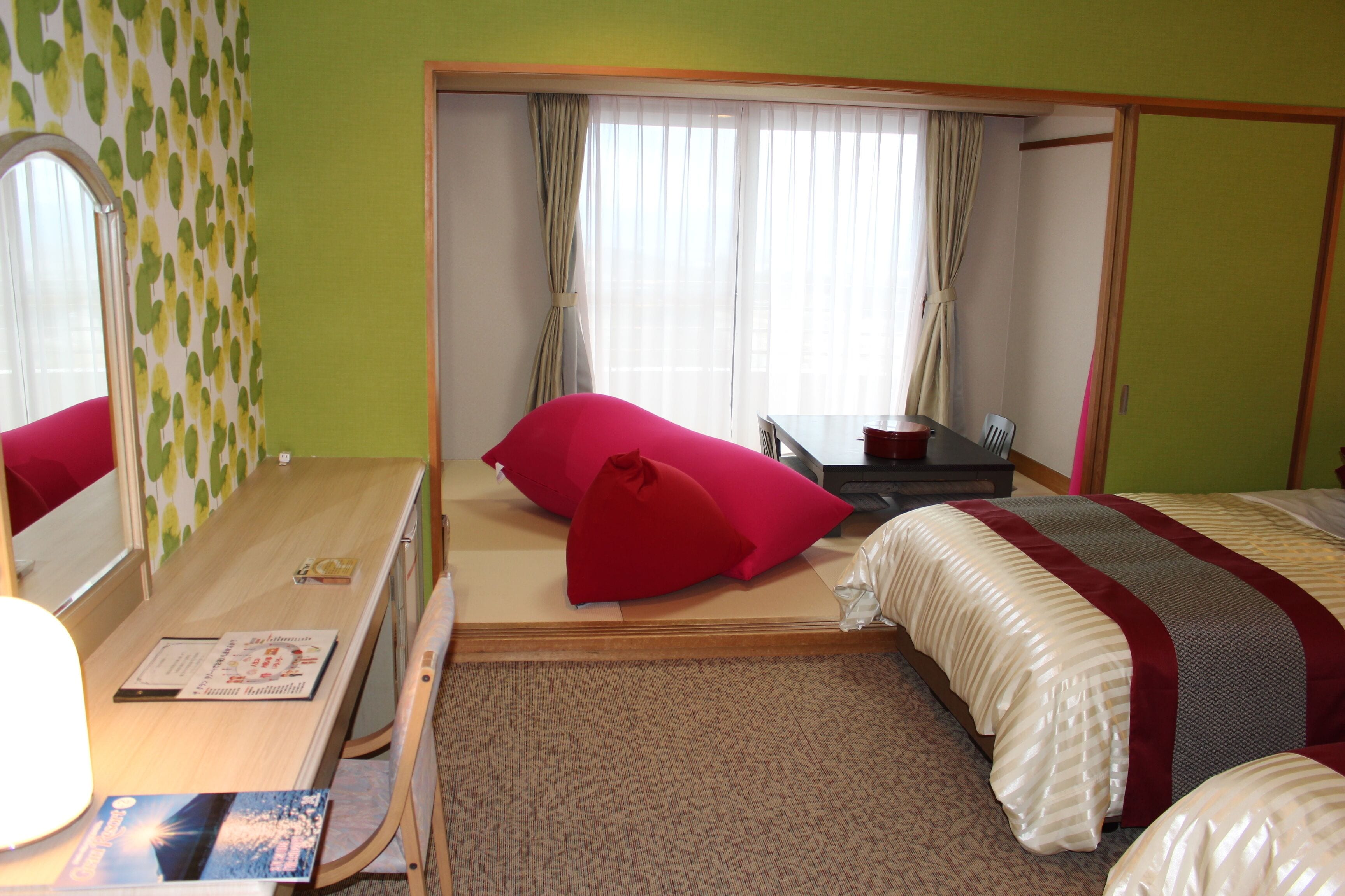 Ein Hotelzimmer mit Bett, Schreibtisch, Stuhl, Spiegel und einem fensterbepflanzten Fenster.