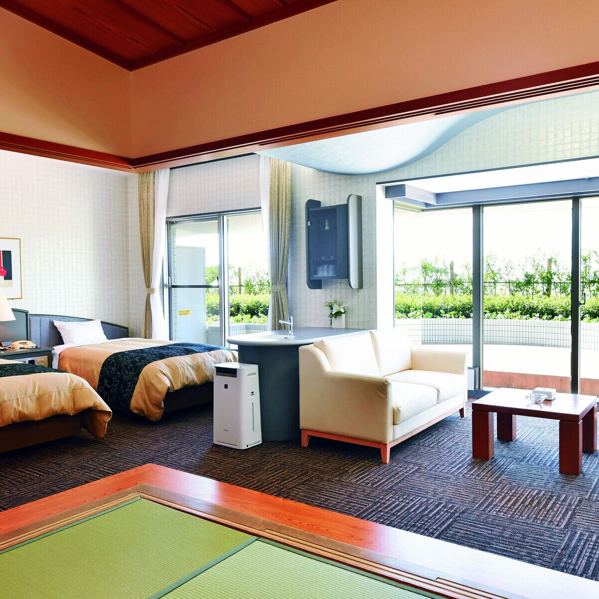 A hotel room with a bed, a desk, a chair, a sofa, a small table, and a large window offering a view of greenery.