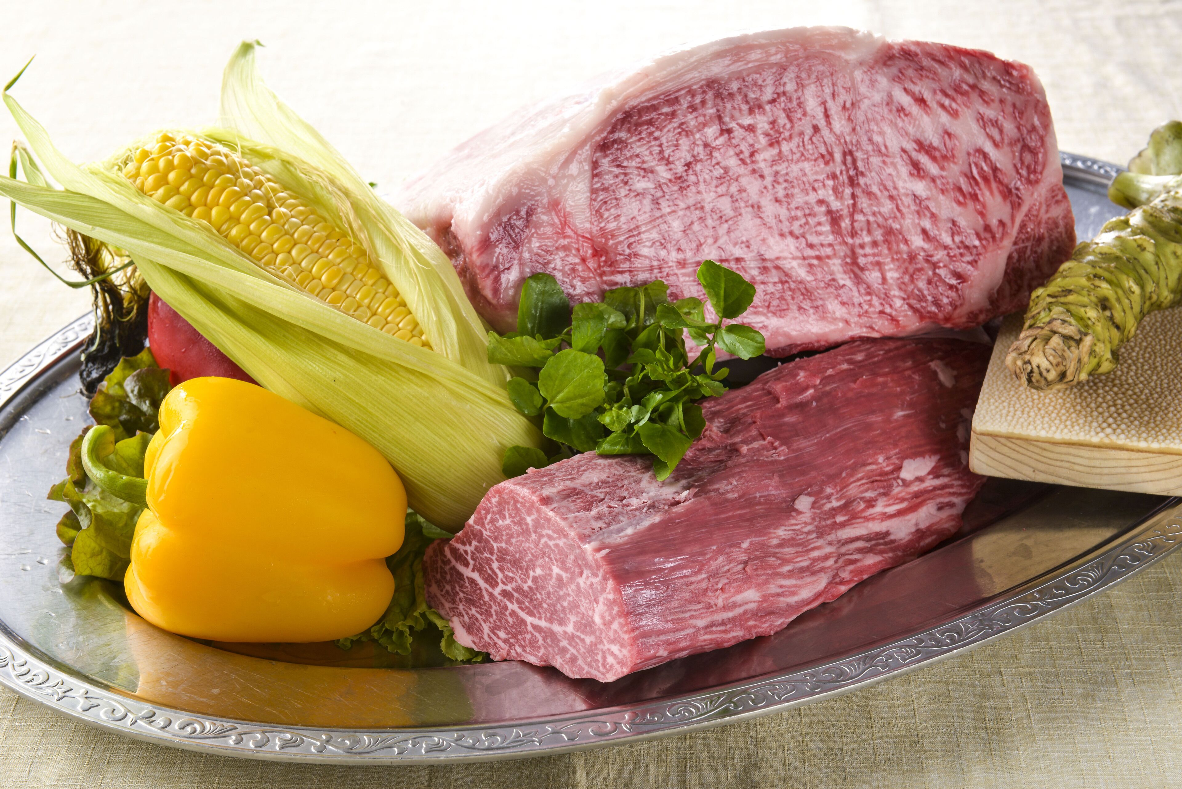 A plate with raw meat, corn on the cob, and a yellow bell pepper.