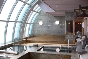 Un espace piscine intérieur moderne, doté de parois en verre transparent, d’un mur en pierre et d’un coin salon avec des bancs.