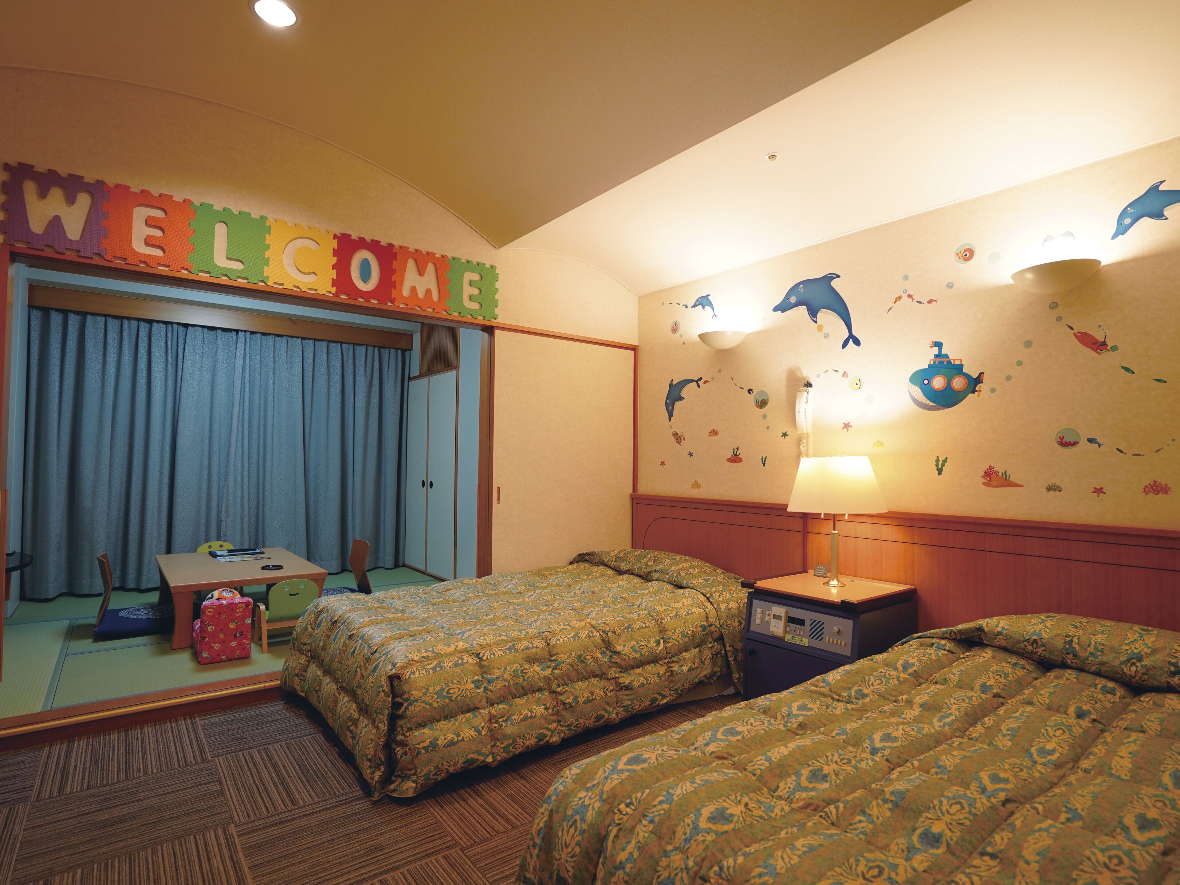 A hotel room with two beds, a desk, and a chair. There is a welcome sign above the door.