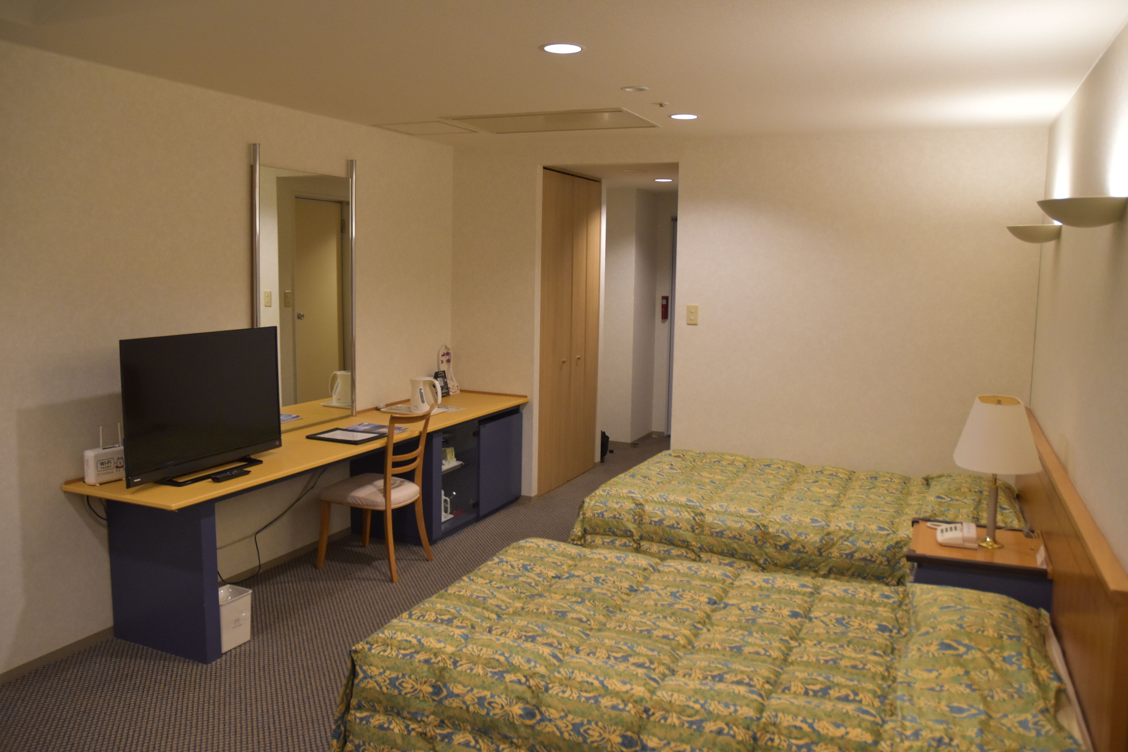 A hotel room with a desk, a television, two beds with matching bedding, a chair, a lamp, and a small cabinet.
