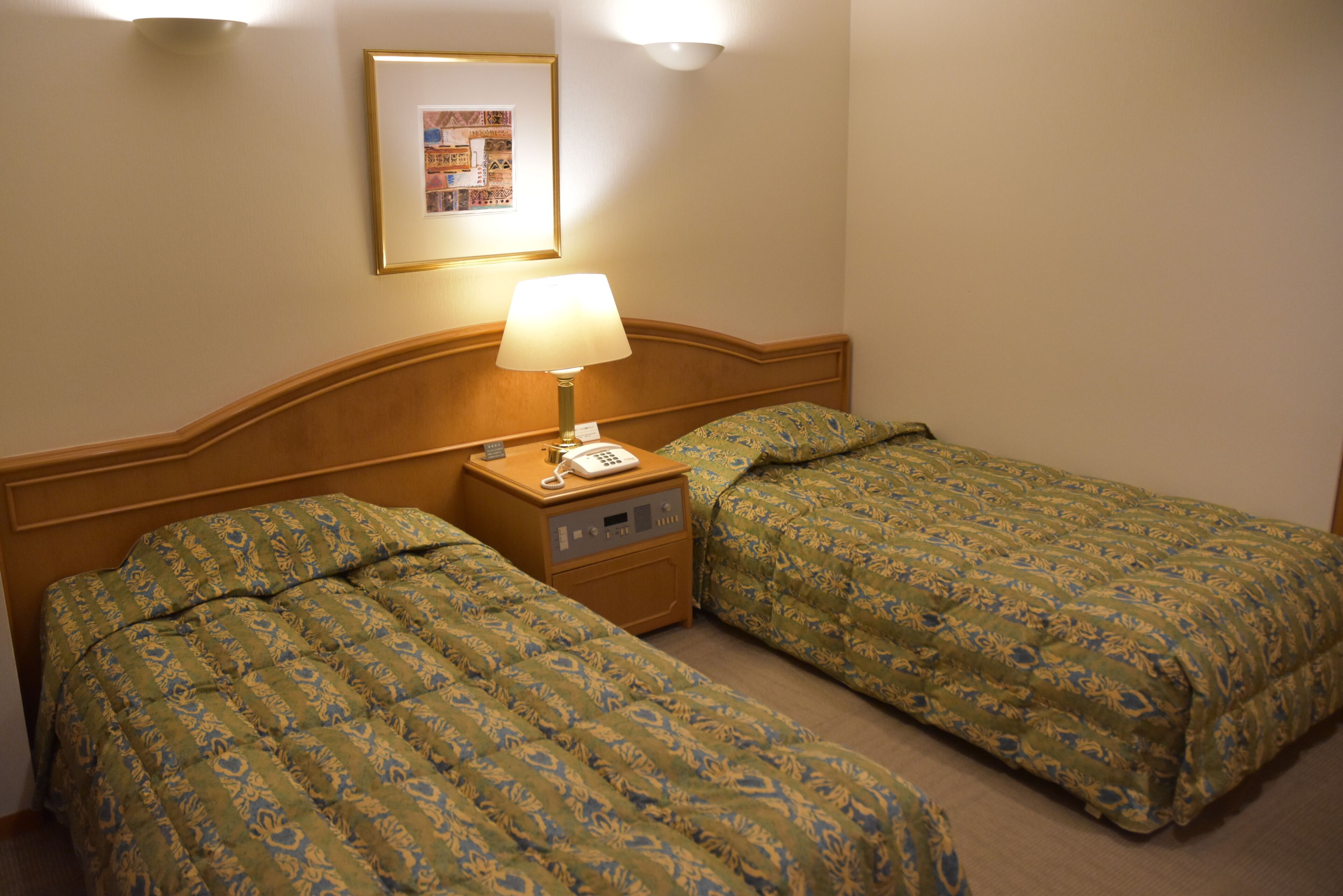 Een hotelkamer met twee enkele bedden, een nachtkastje met een lamp, een telefoon en een lijst met een afbeelding.