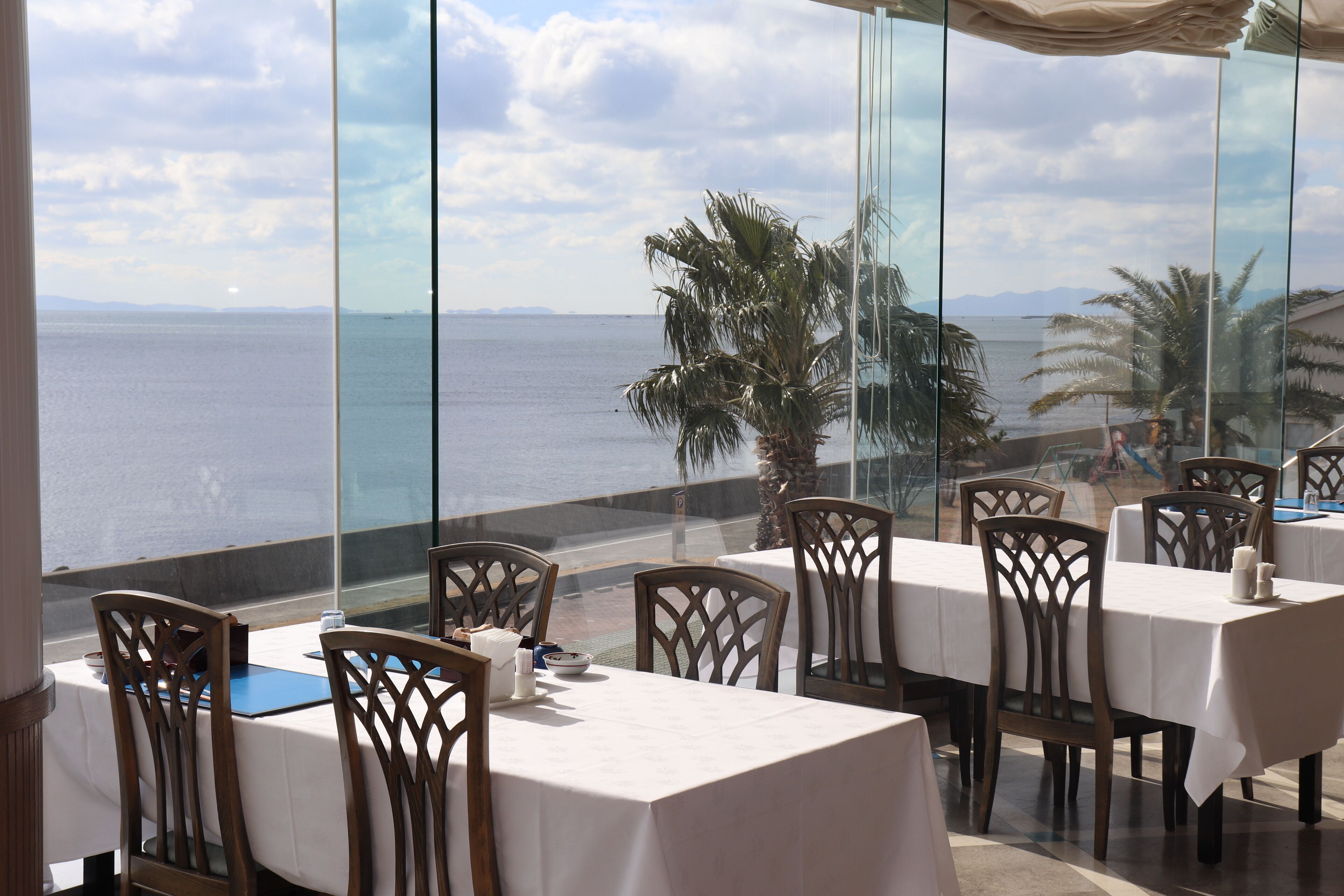 A restaurant with tables and chairs, a view of the sea, and palm trees.