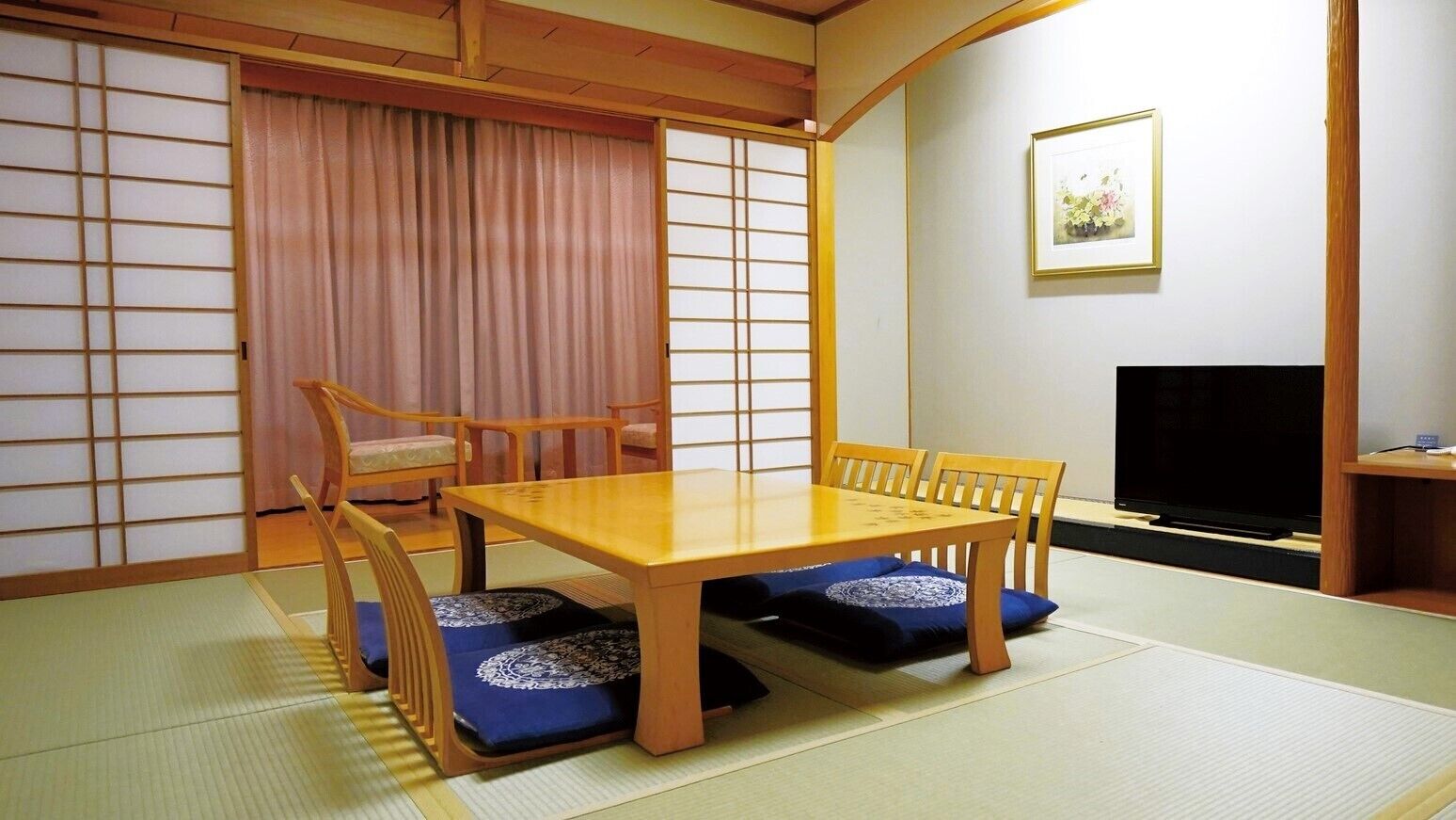 Et traditionelt japansk rum med tatami-gulv, et lavt træbord med puder, et fjernsyn og skydedøre.
