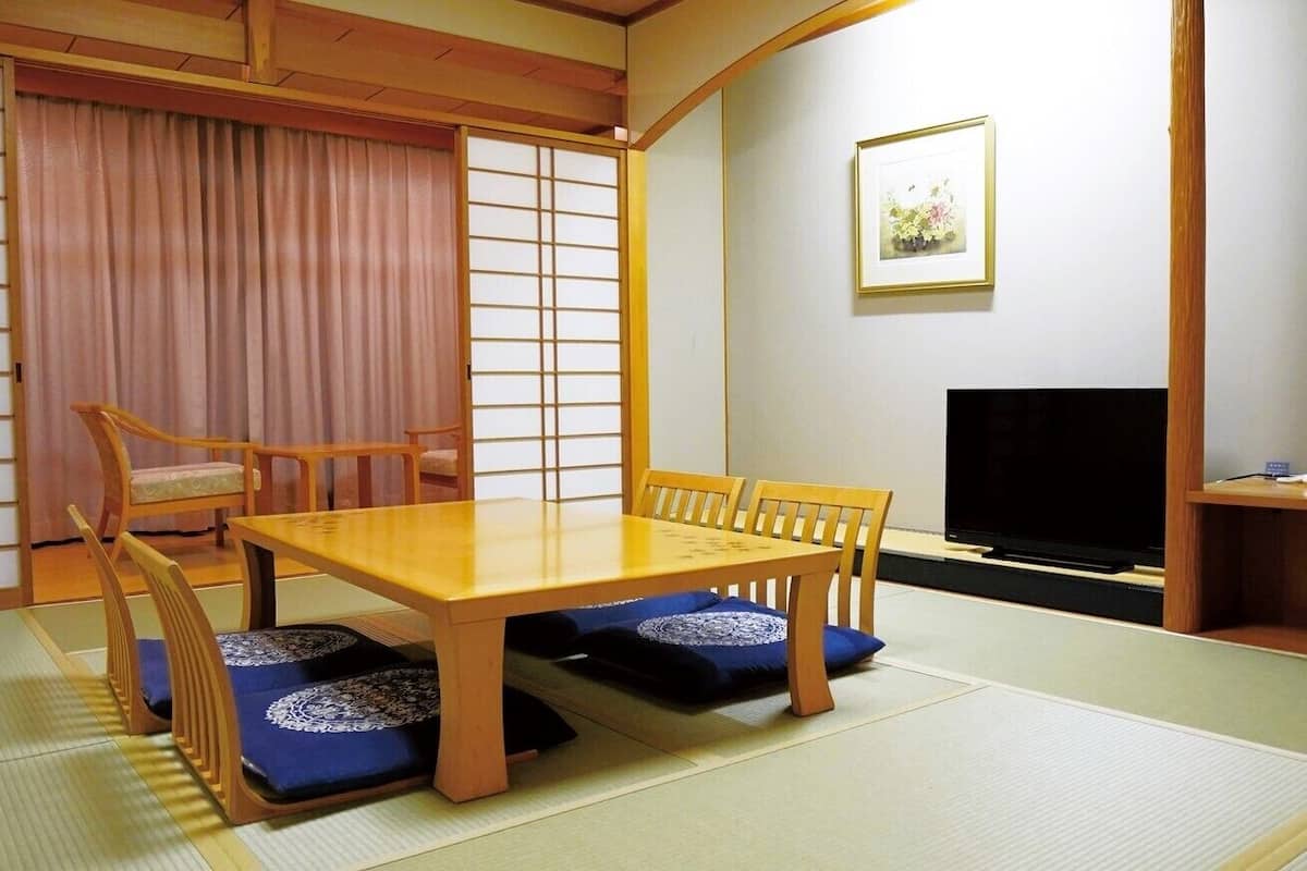 Una habitación japonesa tradicional con suelo de tatami, una mesa de madera baja con cojines, un televisor y puertas correderas.