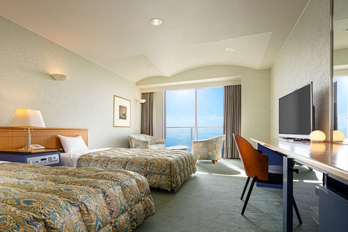Habitación de hotel con dos camas, un escritorio, una silla, un televisor y un ventanal con vistas.