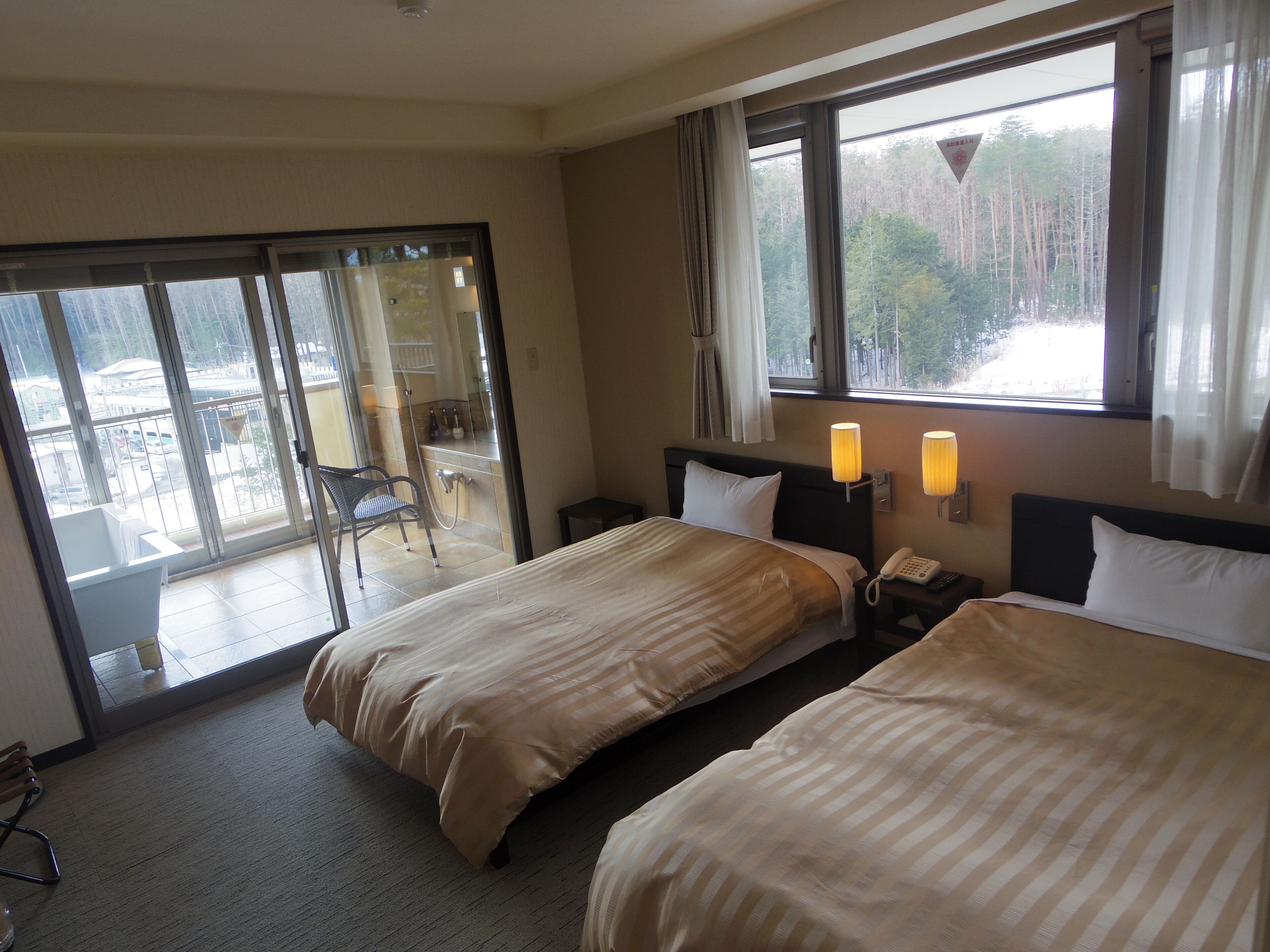 A hotel room with two beds, a bathroom with a shower, and a balcony with a chair.