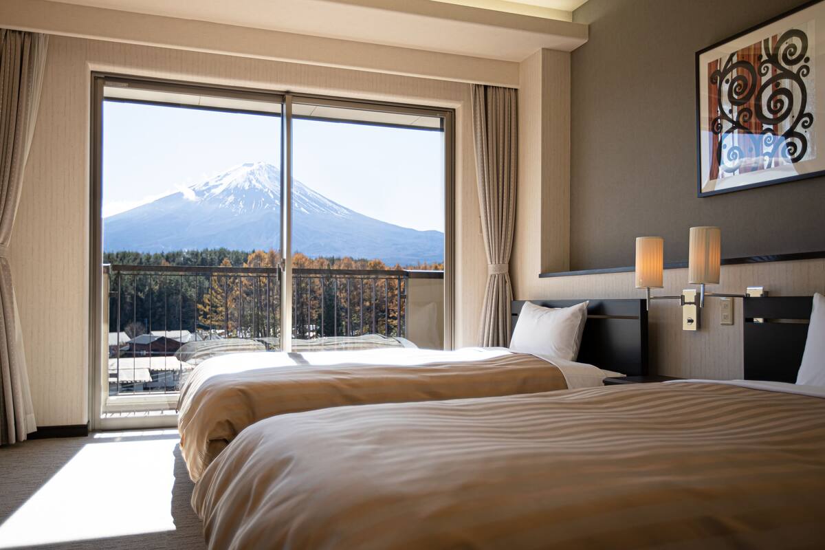 Habitación de hotel con un ventanal que ofrece vistas a una montaña y un bosque. Hay dos camas con ropa de cama color crema, una mesita de noche con una lámpara y una obra de arte colgada en la pared.