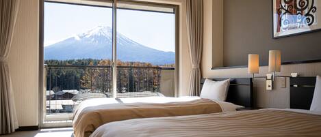 A hotel room with a large window offering a view of a mountain and forest. There are two beds with beige bedding, a nightstand with a lamp, and a wall-mounted artwork.