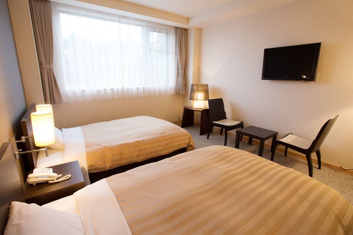 Habitación de hotel con dos camas, un escritorio con teléfono, una silla, una mesa, una lámpara y un televisor montado en la pared.