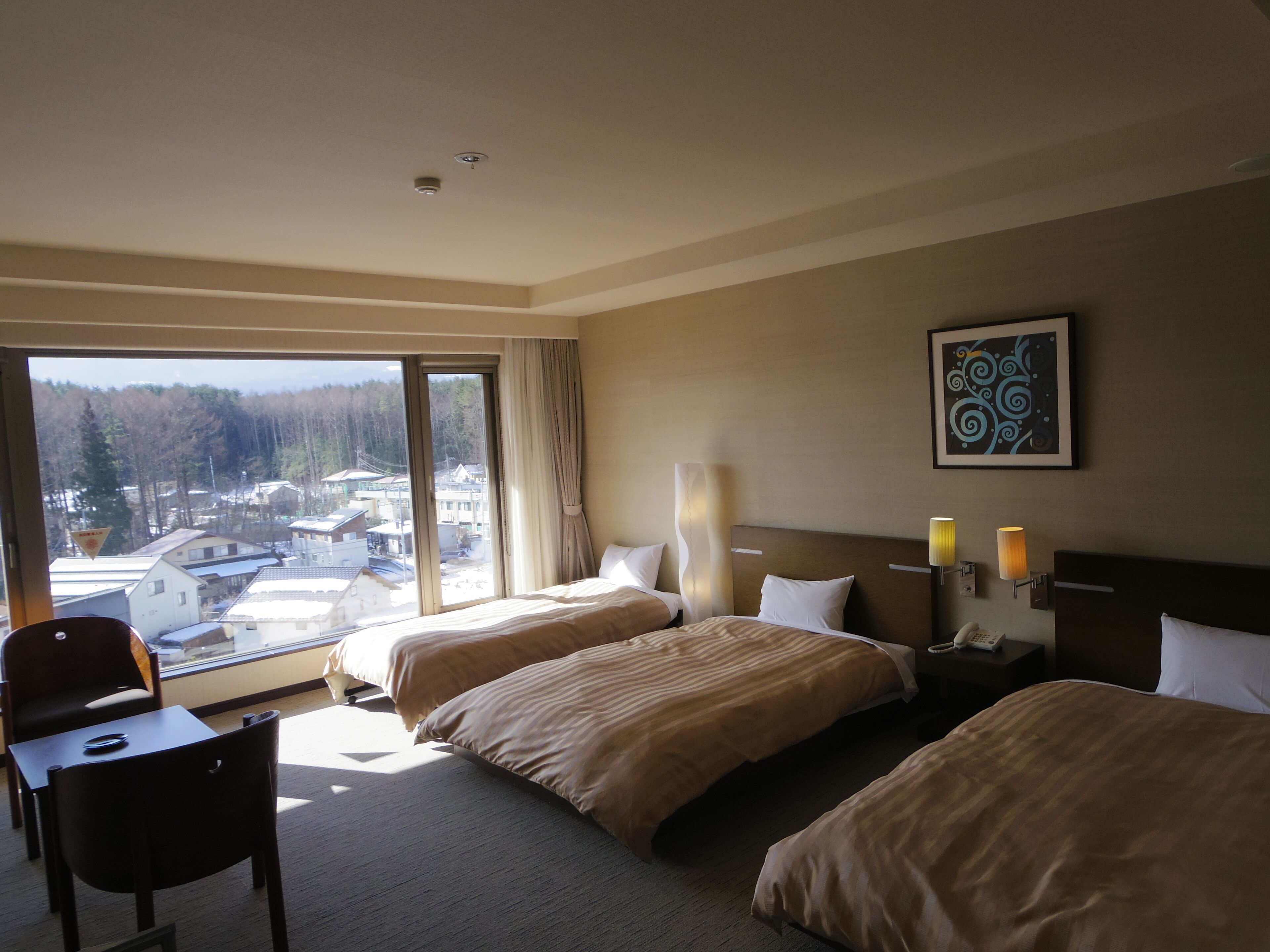 A hotel room with two beds, a chair, a desk, a window with a view of buildings and trees, and a painting on the wall.