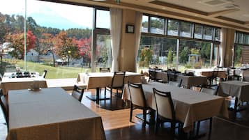 Um restaurante com várias mesas e cadeiras, grandes janelas com vista para árvores e uma montanha.