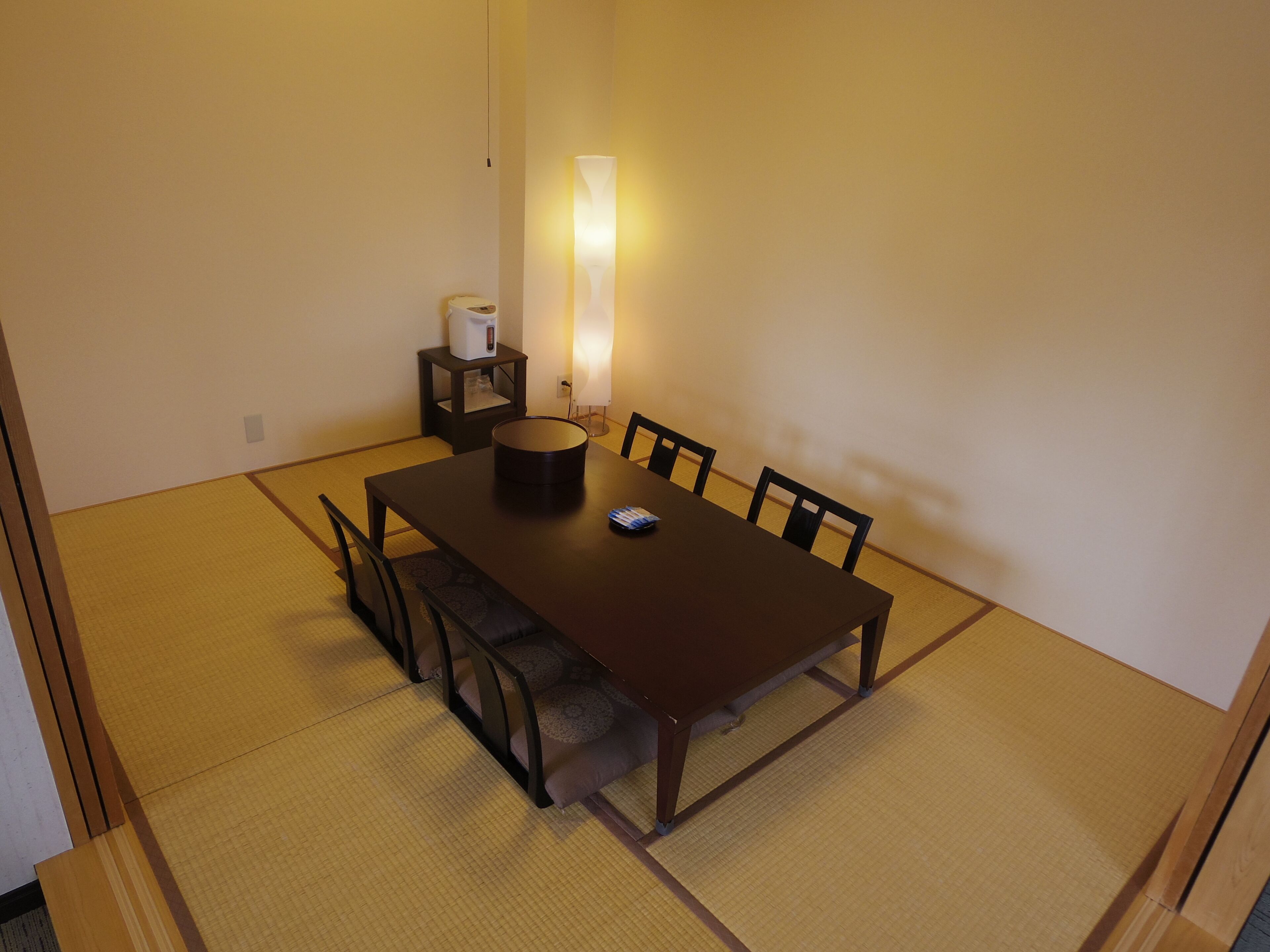A traditional Japanese tatami mat room with a wooden table, chairs, a small side table with a kettle, and a wall-mounted light.