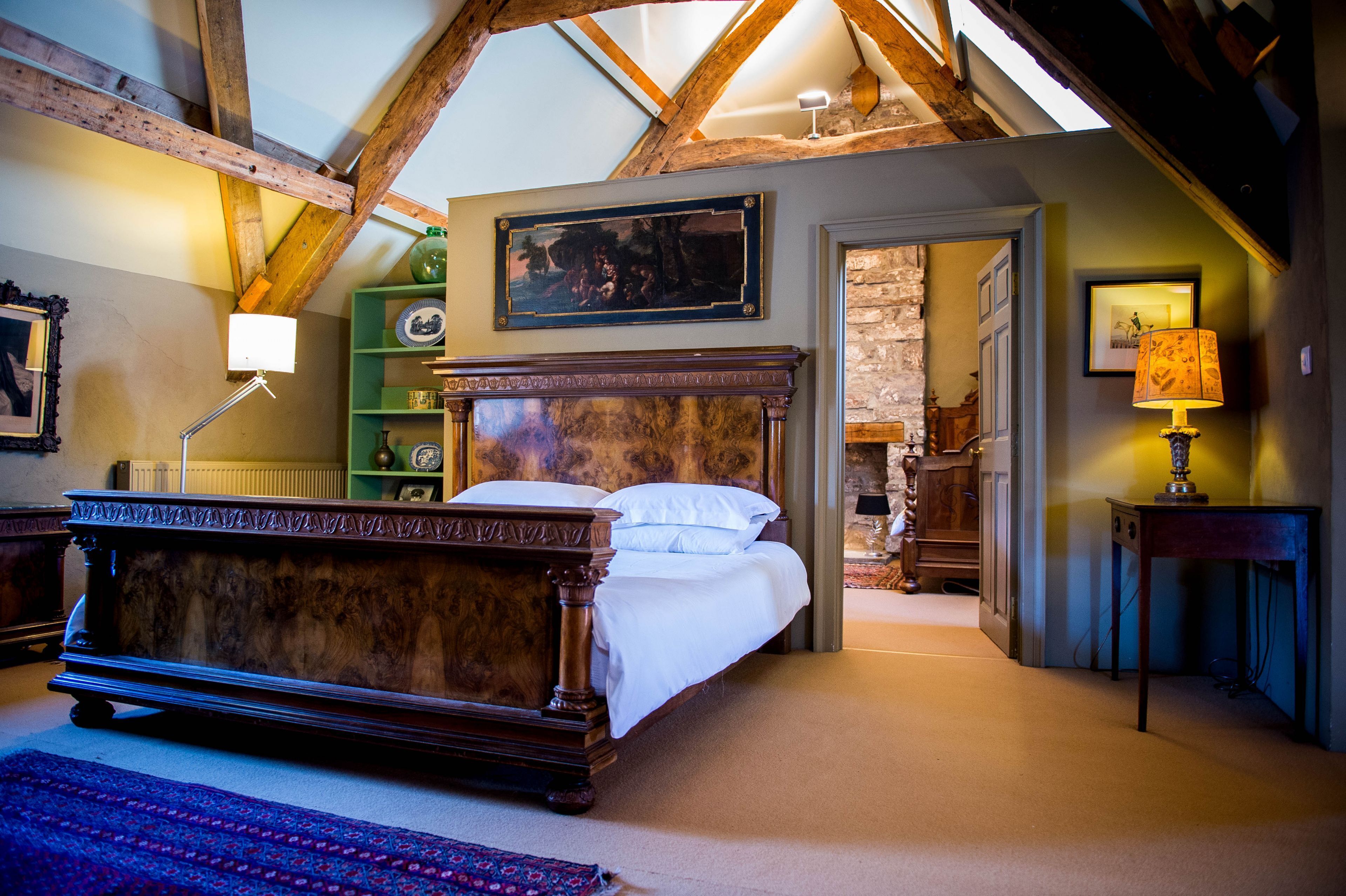 A bedroom with a wooden bed, a green shelf with decorative items, a painting on the wall, and a lamp on a side table.