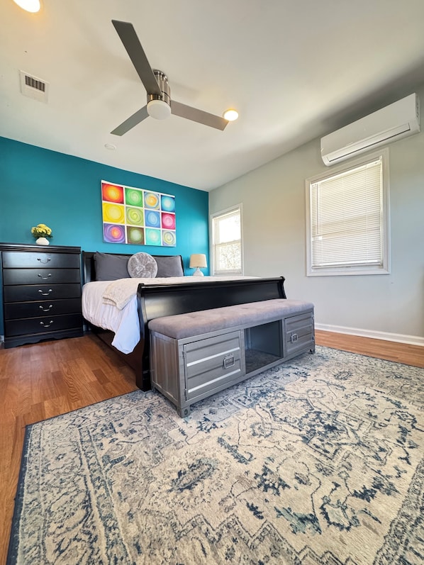 A bedroom with a bed, a ceiling fan, a dresser, a rug, and a wall with colorful artwork.