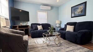 A living room with two dark sofas, a glass coffee table, a patterned rug, a ceiling fan, a flat-screen TV, and an air conditioning unit. - Spring Lake Manor - Rutgers | NYC | Middlesex County New Jersey - EXTENDED STAY (South Plainfield)