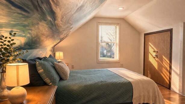 A bedroom with a sloped ceiling, a bed with a quilted cover, a bedside table with a lamp, and a window with blinds. - Spring Lake Manor - Rutgers | NYC | Middlesex County New Jersey - EXTENDED STAY (South Plainfield)