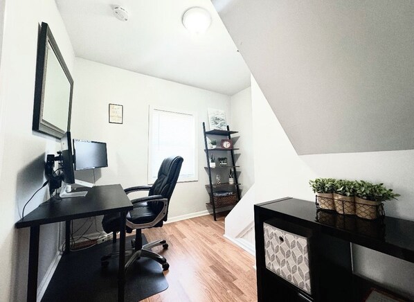 Een werkplek aan huis met een bureau, computer en stoel. Er is een boekenkast met decoratieve objecten en een plant op een plank.