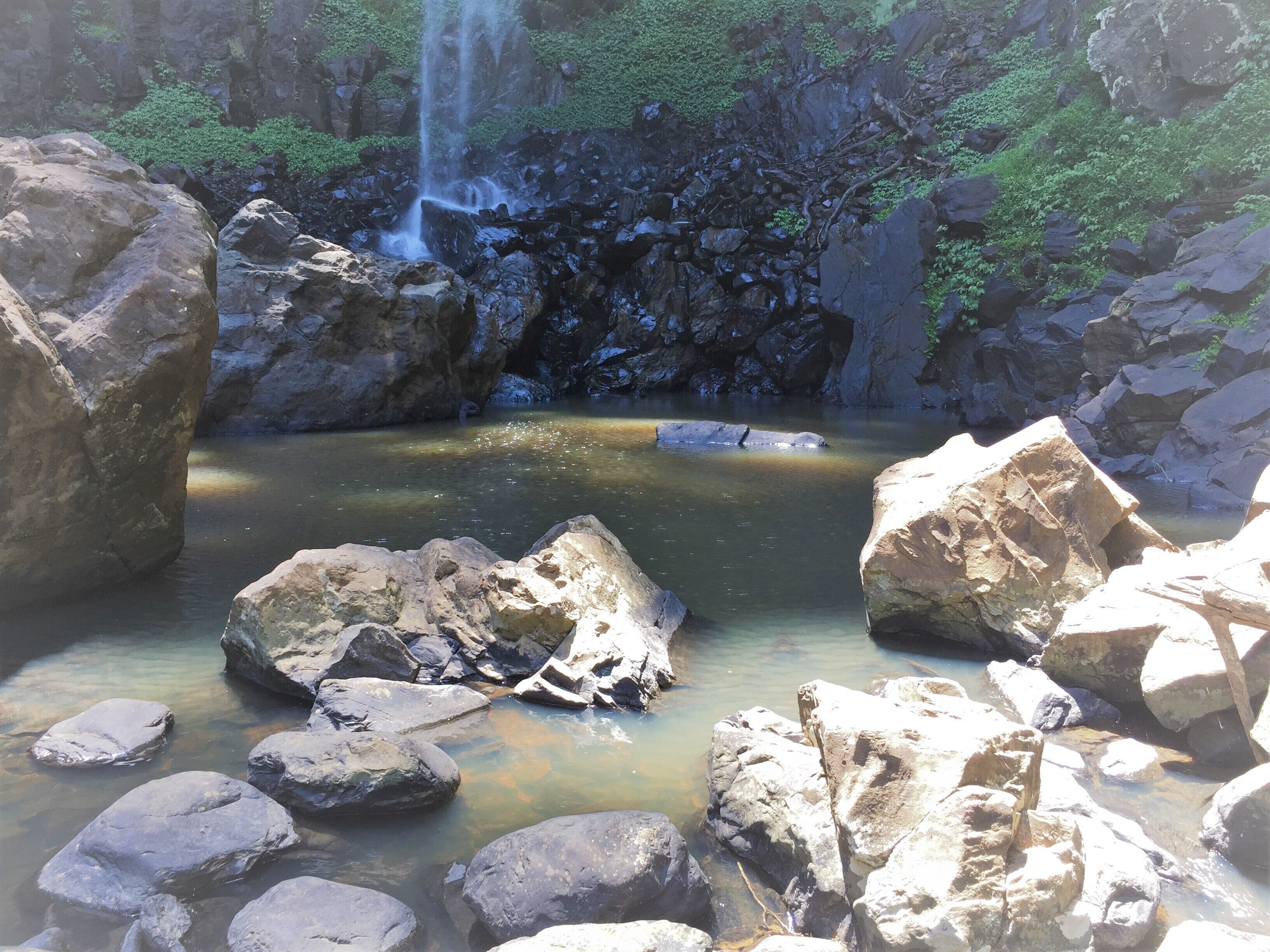 A natural rock pool surrounded by large boulders and a waterfall in the background.