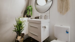 A modern bathroom with a white vanity, a round mirror, a macrame wall hanging, and a potted plant. - Nature Domes (Toms Creek)