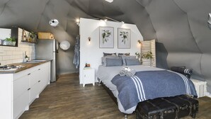 A compact, modern bedroom with a loft-style ceiling, a central bed with a blue blanket, a small bedside table, and a white nightstand. - Nature Domes (Toms Creek)