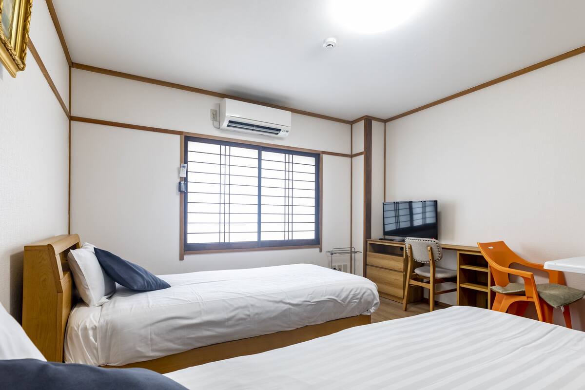 Une chambre avec deux lits, un bureau avec une télévision et une fenêtre avec des stores.