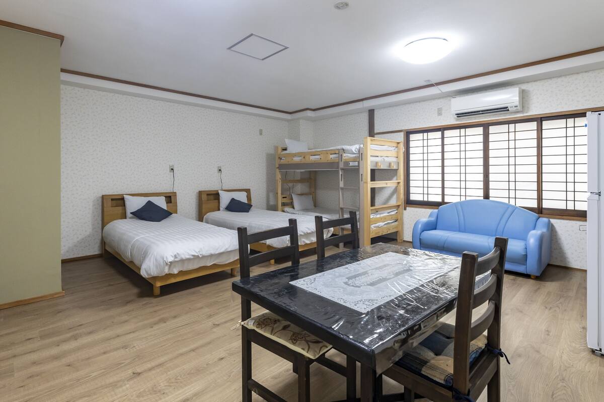 Une chambre avec deux lits simples, un lit superposé, une table à manger avec des chaises, un canapé bleu et un climatiseur fixé au mur.