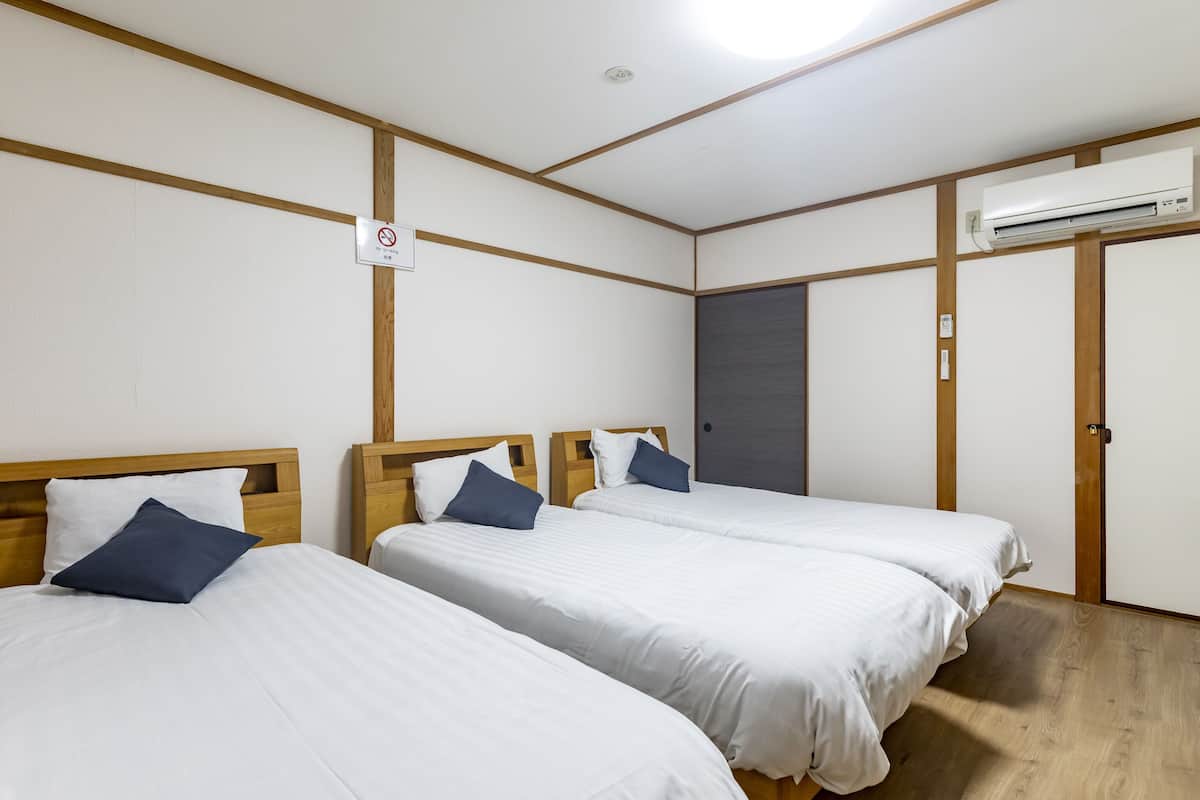 Une chambre d’hôtel avec trois lits simples, des têtes de lit en bois, du linge de lit blanc, un luminaire au plafond, un appareil de climatisation et une porte avec une serrure.