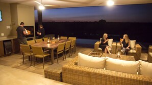 Un área de comedor exterior moderna con una gran mesa de madera, mobiliario de mimbre y vistas al cielo nocturno.