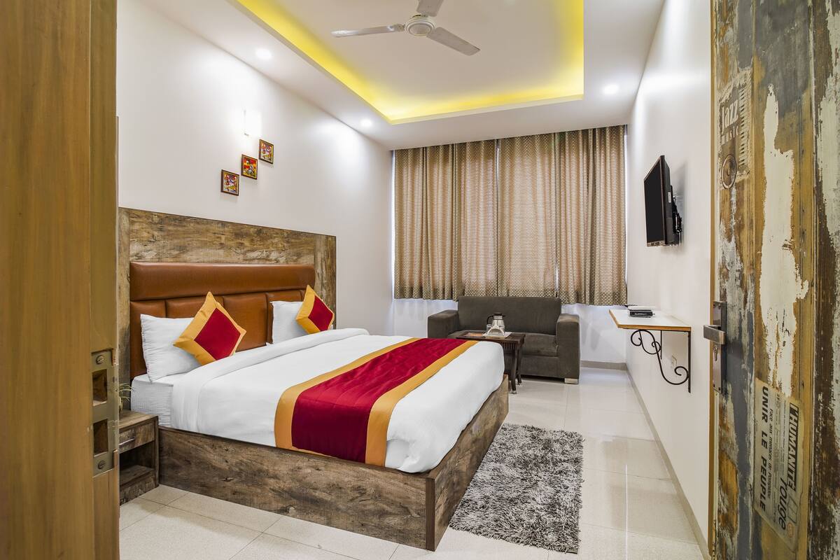 Une chambre d’hôtel avec une tête de lit en bois, un grand lit avec une literie blanche agrémentée de touches de rouge et d’or, un petit bureau avec une chaise, un canapé et une grande fenêtre avec des rideaux.