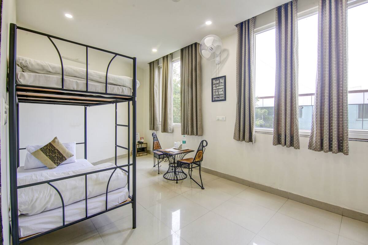 Une chambre avec un lit superposé, une table avec deux chaises, un ventilateur et des rideaux aux fenêtres.