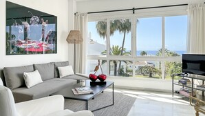 A modern living room with a grey sofa, a coffee table with decorative items, a TV on a stand, and a large window with a view of palm trees and buildings. - J07 Jardines 4 - Apartment with sea views (Sitio de Calahonda)
